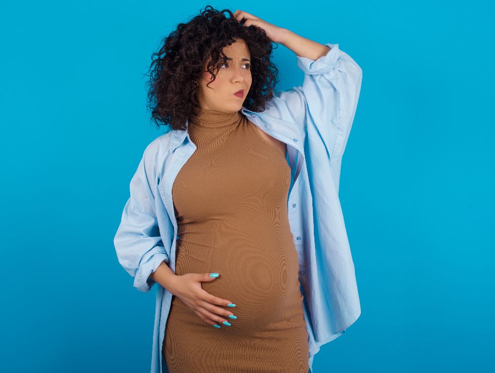 young Arab pregnant woman wearing dress against blue wall confuse and wonder about question. Uncertain with doubt, thinking with hand on head. Pensive concept
